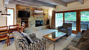 A spacious living room with a stone fireplace, comfortable seating, a flat-screen TV, and large windows offering a view of the outdoors. - 🌲Timberline A2H Deluxe 2-Bedroom ~Hot Tub~Pool~WiFi~Ski-in/out~Local Shuttle🌲 (Snowmass Village)