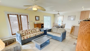 A spacious living area with a sofa, a coffee table, and a dining table with chairs. There is a ceiling fan, a window with blinds, and a kitchen area in the background.