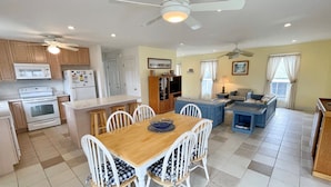 A compact kitchen with a stove, microwave, and refrigerator. There is a dining area with a wooden table and white chairs. The room has a ceiling fan and a window with curtains.