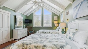 Une chambre avec un plafond en pente, un grand lit avec une literie à motifs floraux, une commode, une chaise et un téléviseur fixé au mur.