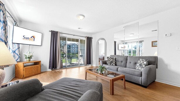 Moderni olohuone, jossa on harmaa sohva, puinen sohvapöytä ja litteänäyttöinen televisio, jolla näkyy ohjelma nimeltä ”Sunset Nightly Rentals”.