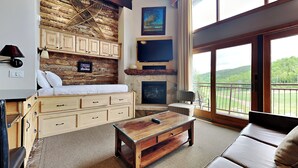 A cozy room with a stone fireplace, a bed with white linens, a wooden coffee table, and a view of a green landscape through large windows. - 🌲Timberline 317 Studio Loft Deluxe ~ Hot Tub~Pool~WiFi~Ski-in/out~Local Shuttle🌲 (Snowmass Village)
