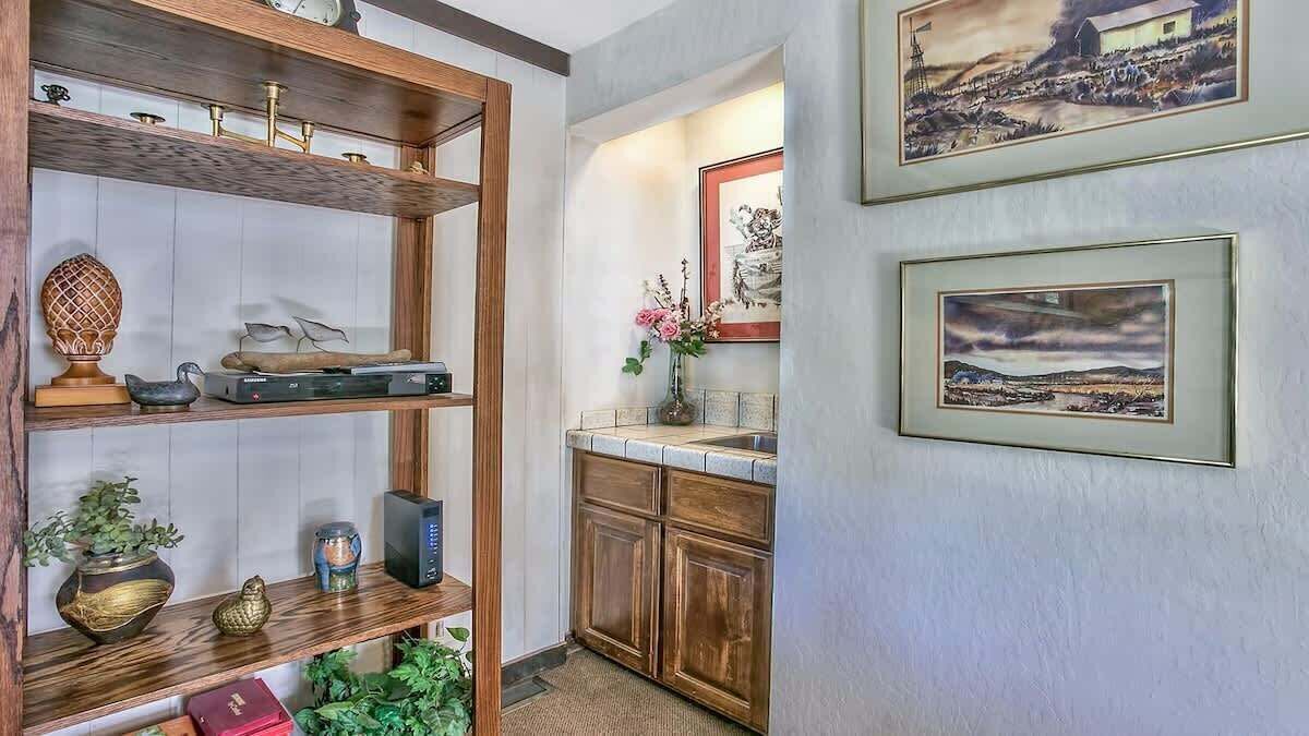 A room with a wooden shelf holding decorative items, a small table with a vase of flowers, and a framed picture on the wall.