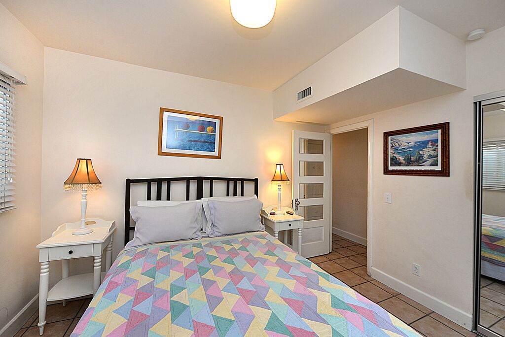 A bedroom with a bed, two bedside tables with lamps, a door leading to another room, and two framed pictures on the walls.