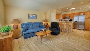 A living room with a sofa, a coffee table, and a chair. There is a kitchen area with wooden cabinets and a refrigerator.