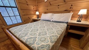 Ein Schlafzimmer einer Holzhütte mit einem Bett, zwei Nachttischen mit Lampen und einem Fenster mit Jalousien.