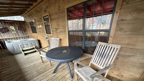 Eine Holzterrasse mit einem runden Tisch, zwei Stühlen, einem Whirlpool und einem Fenster mit rotem Vorhang.