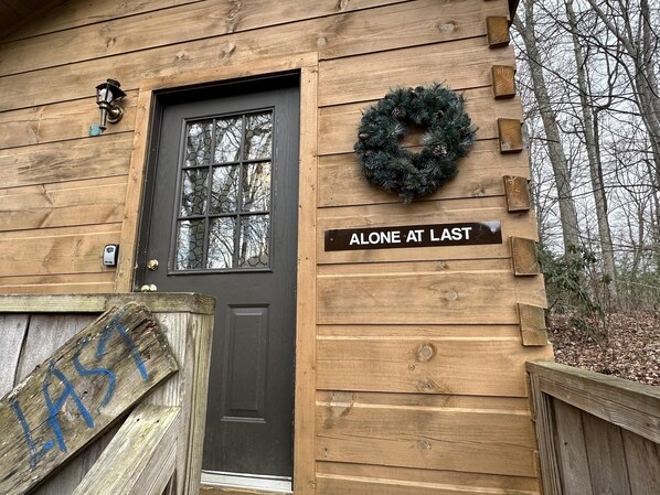A wooden cabin with a black door, a stone-framed window, a wreath, and a sign reading 'ALONE AT LAST'. - Alone at last! Hiking, hot tub, grill, view. (Asheville)