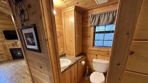 Ein Badezimmer einer Holzhütte mit einem Waschbecken auf einer Granittheke, einer Toilette und einem Fenster mit einem Karomuster-Vorhang.