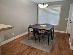 Uma área de refeições com uma mesa e cadeiras, um tapete, uma janela com persianas e uma porta.