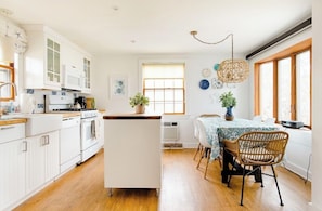 A modern kitchen with white cabinetry, a central island, and a dining area with wicker chairs and a table. - Starfish Cottage★0.4mi walk Beach & Village★Pet Friendly★Bikes★Fireplace★Firepit (Dennis)