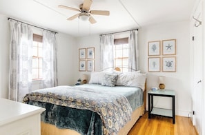 A bedroom with a bed, a ceiling fan, a bedside table with a lamp, and framed pictures on the wall. - Starfish Cottage★0.4mi walk Beach & Village★Pet Friendly★Bikes★Fireplace★Firepit (Dennis)