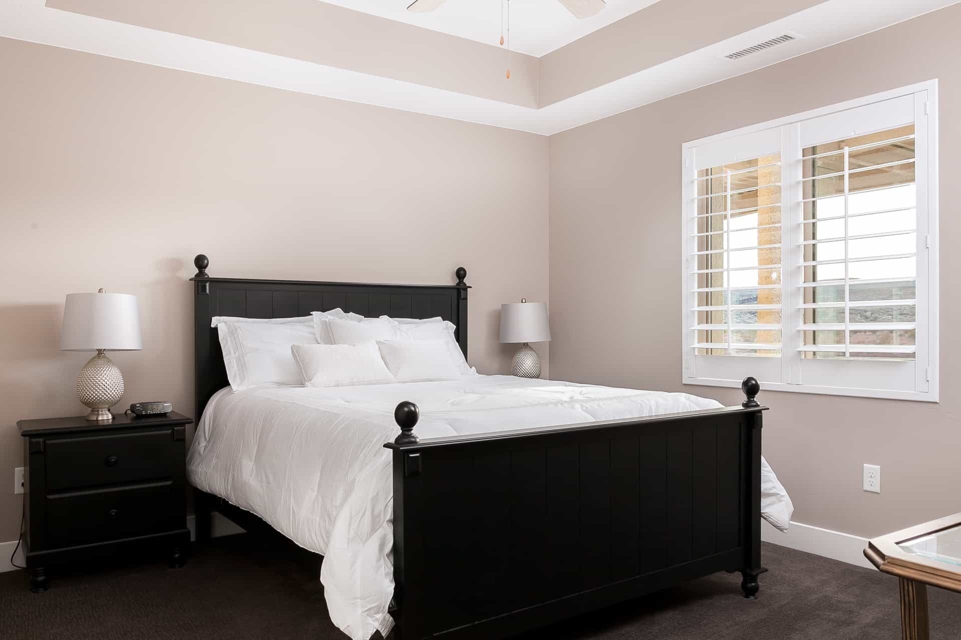 A bedroom with a black four-poster bed, two bedside tables with lamps, a window with blinds, and a carpeted floor.