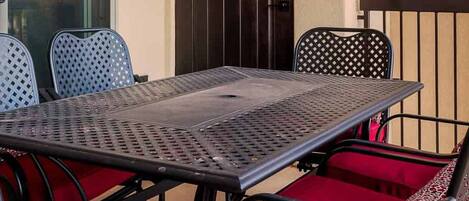 Un balcón con una mesa y sillas de metal, una puerta corredera de vidrio y una puerta oscura.