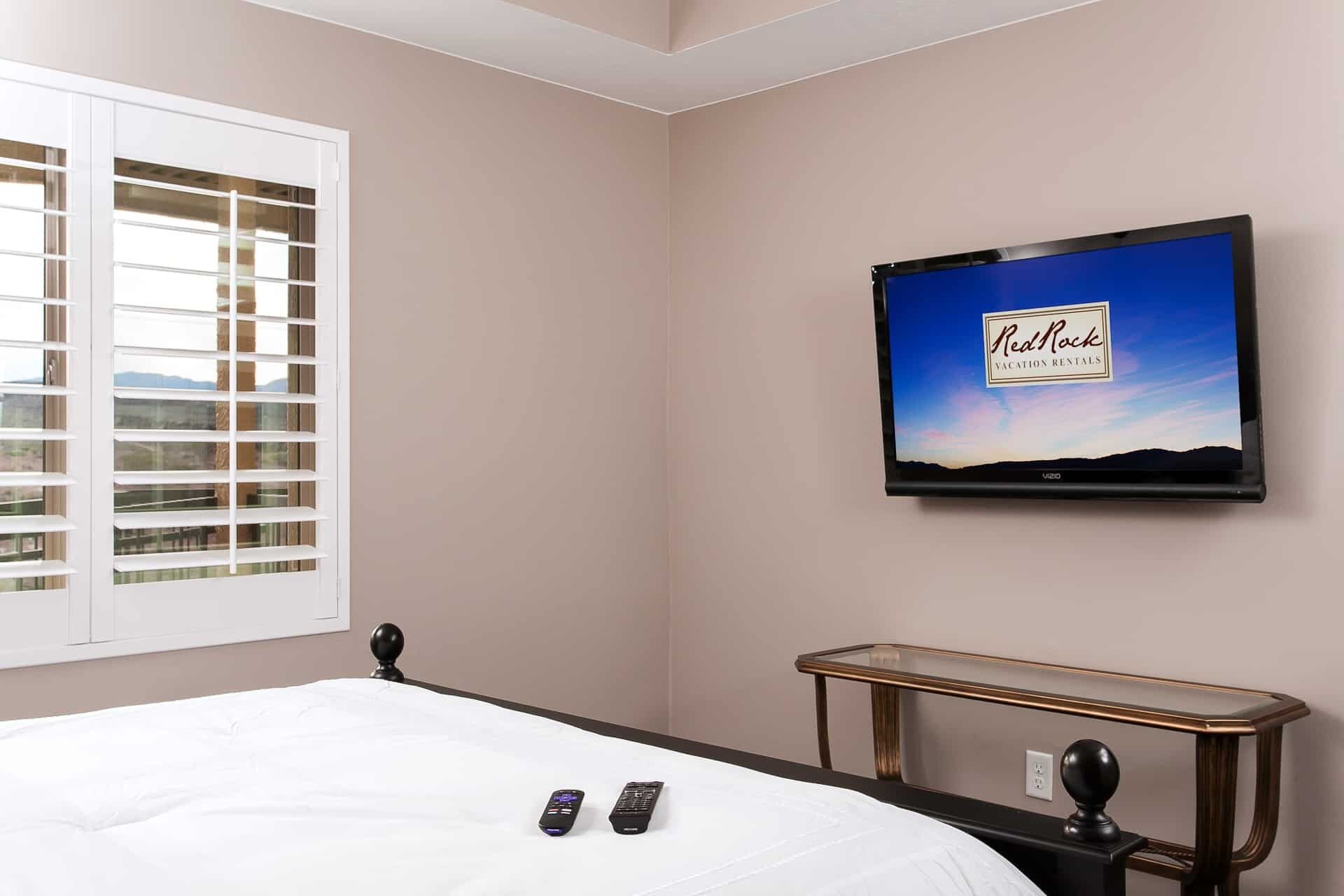 Ein Hotelzimmer mit einem Bett, einem an der Wand montierten Fernseher, der 'RedRock Vacation Rentals' anzeigt, und einem Nachttisch mit einer Fernbedienung.