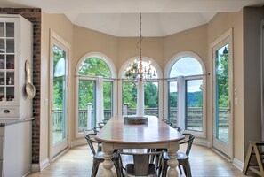 A dining room with a large table, chairs, and a chandelier. There are large windows and a view of greenery outside.