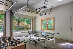 A ping pong table with a table tennis net, a foosball table, and a window with a view of greenery.