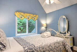 A bedroom with a bed, a window with a floral curtain, a bedside lamp, and a mirror on a dresser.