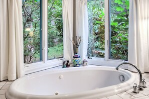 A large white bathtub with a modern faucet, positioned in front of a window with white curtains, overlooking a lush green garden.