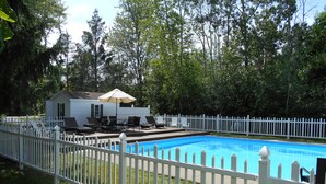 Ein Swimmingpool, umgeben von einem weißen Zaun mit kleinen Haus und Gartenmöbeln im Hintergrund.
