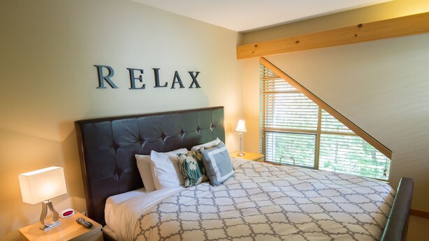 Quarto com cama, mesa de cabeceira, luminária, relógio e janela com persianas.