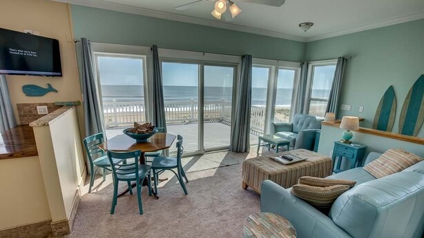 A spacious living room with a large sliding glass door leading to a balcony overlooking the ocean. There is a dining area with a table and chairs, a sofa, and a television mounted on the wall. - Pointe O'View | Oceanfront | Private Pool, Hot Tub (Virginia Beach)
