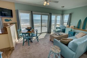 A spacious living room with a large sliding glass door leading to a balcony overlooking the ocean. There is a dining area with a table and chairs, a sofa, and a television mounted on the wall. - Pointe O'View | Oceanfront | Private Pool, Hot Tub (Virginia Beach)
