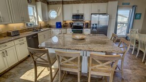 A kitchen with white cabinets, a granite countertop, a stainless steel refrigerator, and wooden chairs. - Pointe O'View | Oceanfront | Private Pool, Hot Tub (Virginia Beach)