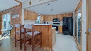 A kitchen with wooden cabinets, a black refrigerator, and a microwave. There is a small island with stools and a table. - 1523 - Moby Vic's - Whalehead Retreat, 200 Yards to Corolla Beach (Corolla)