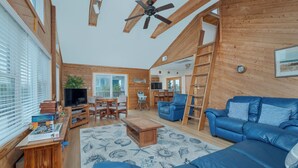 A spacious living room with a blue sofa, wooden ceiling beams, a ceiling fan, and a television. - 1523 - Moby Vic's - Whalehead Retreat, 200 Yards to Corolla Beach (Corolla)