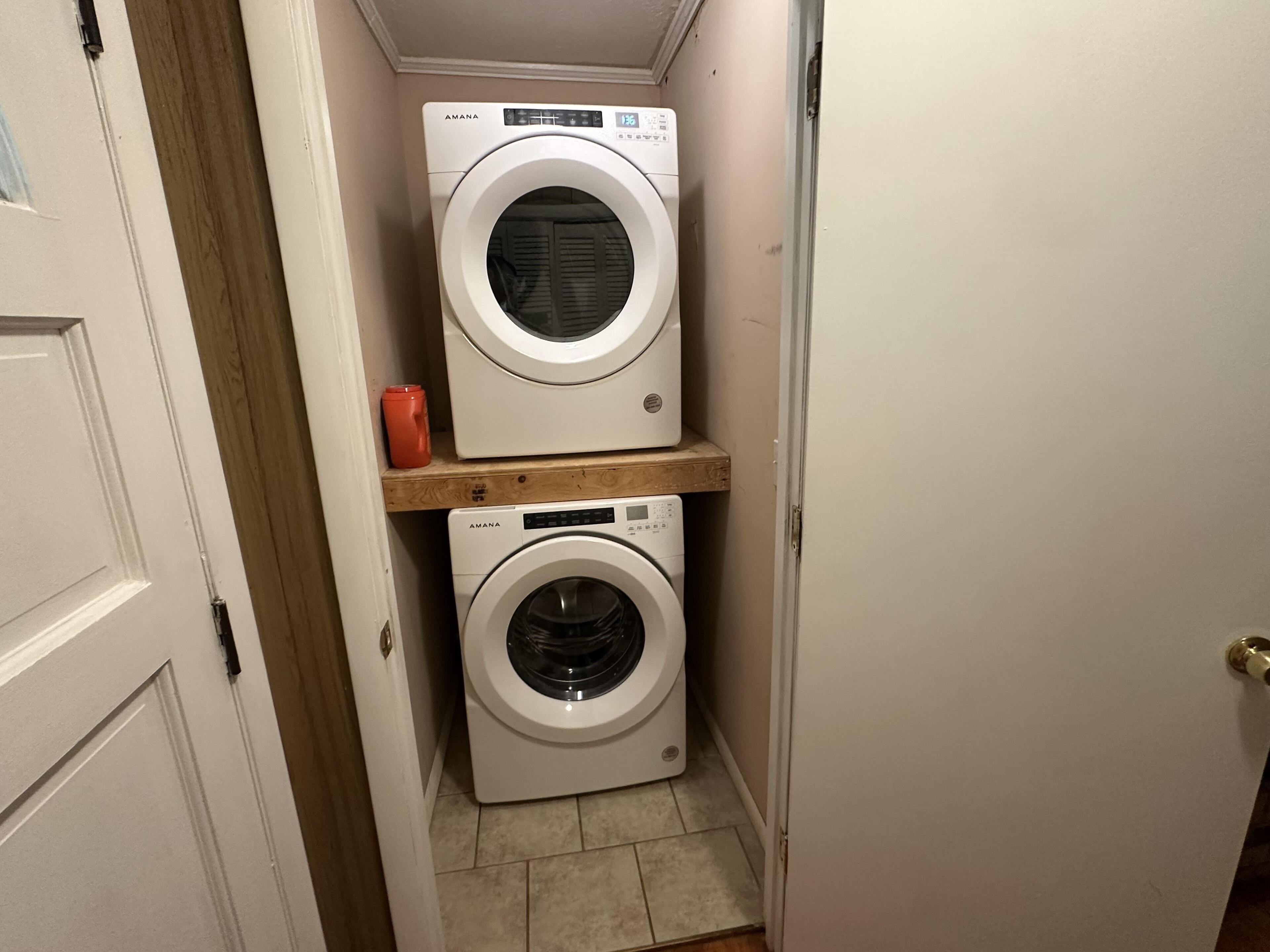 A laundry room with two white washing machines, one on top of the other, and a red container on a wooden shelf.