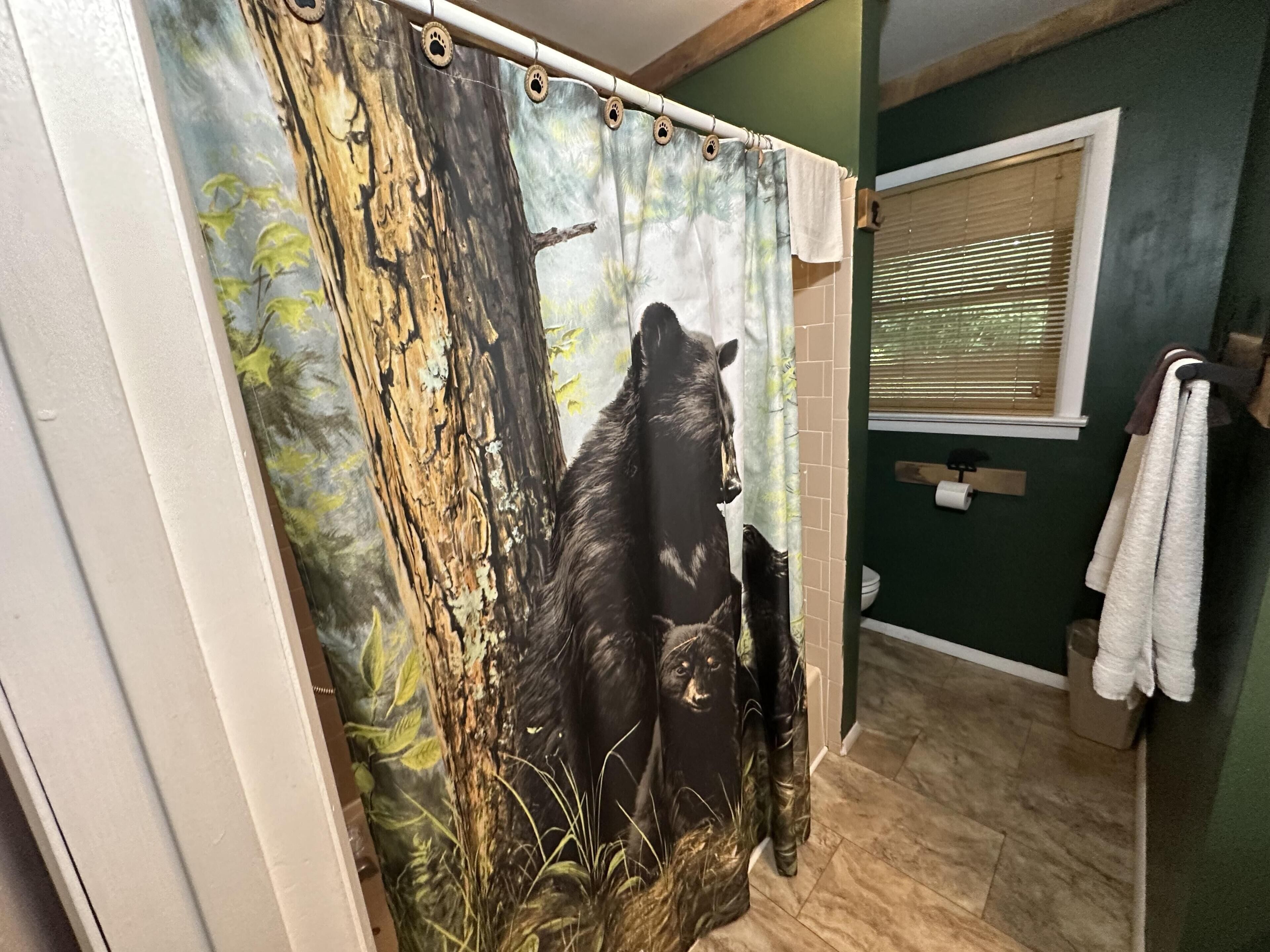 A bathroom with a forest-themed shower curtain, a window with blinds, and a towel rack with towels.