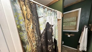 Ein Badezimmer mit Duschvorhang mit Waldbild, einem Fenster mit Jalousien und einem Handtuchhalter mit Handtüchern.