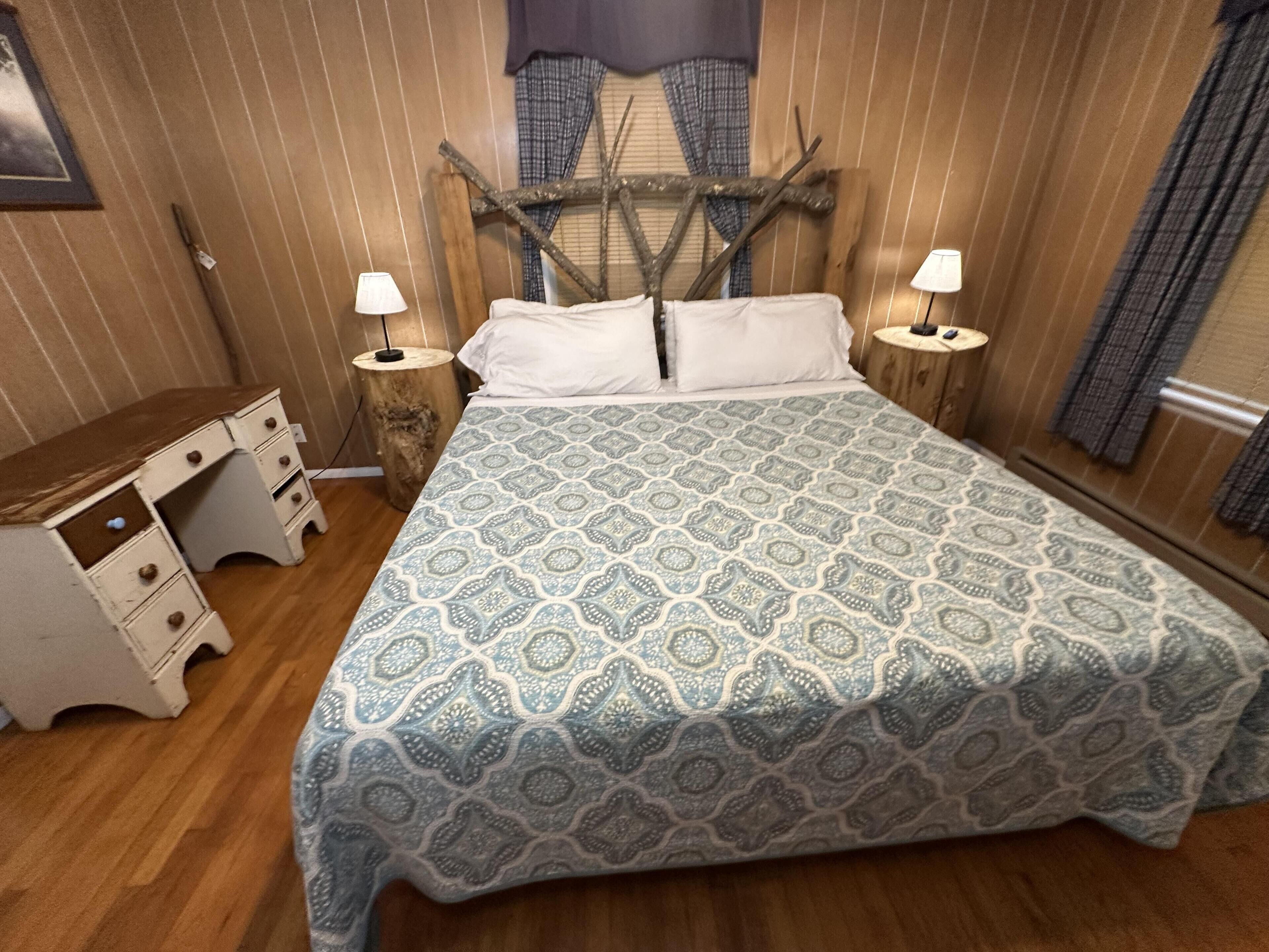 Une chambre à coucher avec une couvre-lit à motifs, un bureau en bois, deux lampes de chevet et une tête de lit rustique.