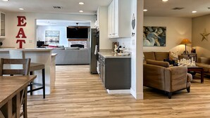 Eine moderne Küche mit einer Kochinsel, ein Essbereich mit Holztisch und -stühlen sowie ein Wohnzimmer mit Sofa und Schachbrett.