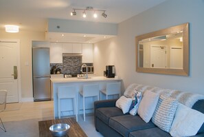 A modern kitchen with white cabinets, a stainless steel refrigerator, and a microwave. There is a dining area with a bar stool and a sofa with decorative pillows.