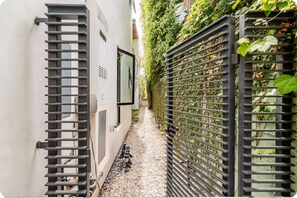 A narrow pathway flanked by a white building and a black metal fence with climbing plants.