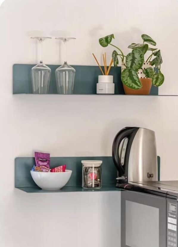 A kitchen area with a microwave, a kettle, a bowl with snacks, and a potted plant on a shelf.