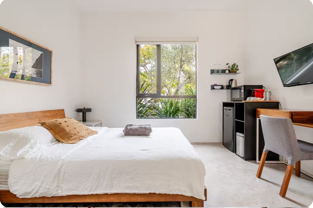 Une chambre à coucher comprenant un lit, un bureau, une chaise, un téléviseur, une fenêtre donnant sur des arbres et un tableau encadré accroché au mur.