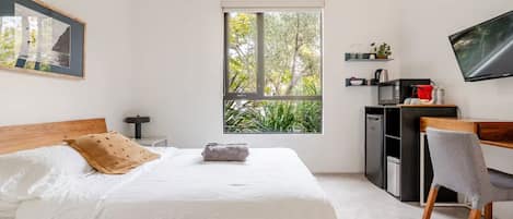 Une chambre à coucher comprenant un lit, un bureau, une chaise, un téléviseur, une fenêtre donnant sur des arbres et un tableau encadré accroché au mur.