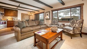 A spacious living room with a brown leather sofa, a wooden coffee table, and patterned armchairs. A kitchen is visible in the background with wooden cabinets and a stone backsplash. - 2 Bed / 2 Bath With Amazing Views Steps From Gondola (Vail)