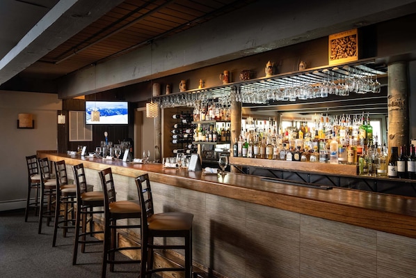 A bar area with a long counter, multiple bar stools, a television screen, and a variety of bottles and glasses.