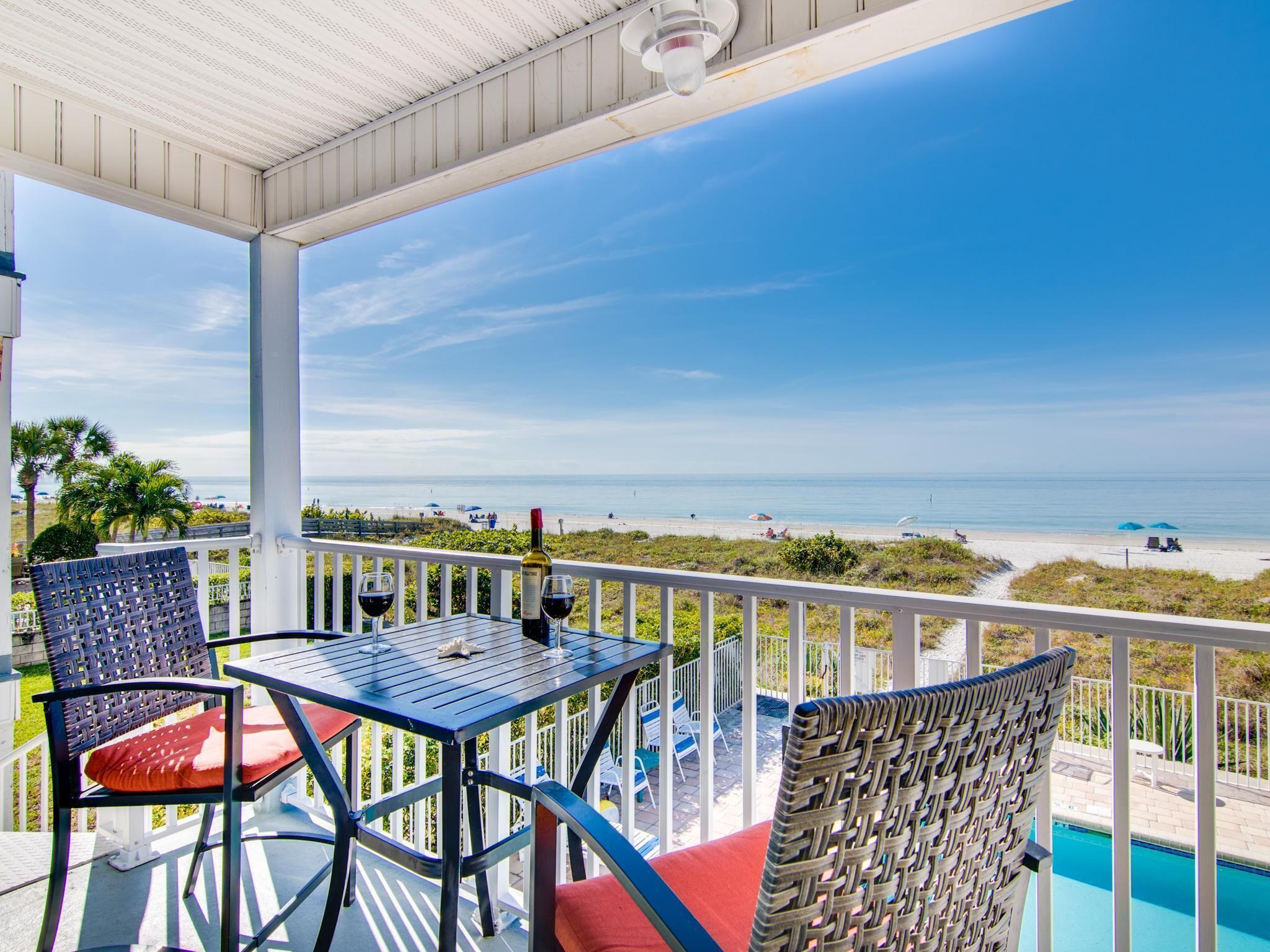 A covered outdoor area with a table and chairs, overlooking a beach and a pool.