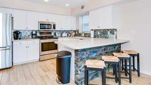 Una cocina moderna con armarios blancos, electrodomésticos de acero inoxidable, una isla grande con taburetes altos y un cubo de basura.