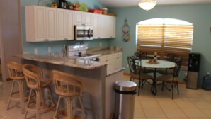 Una cocina con armarios blancos, una isla central con taburetes, una mesa de comedor con sillas y un cubo de basura.