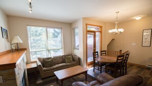 A cozy living room with a sofa, coffee table, and dining table. There is a window with blinds, a lamp, and a door leading outside.