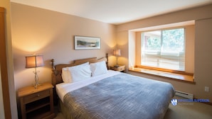 A bedroom with a bed, two bedside tables with lamps, a window with blinds, and a wall-mounted picture.