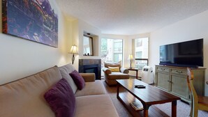 A living room with a sofa, armchair, coffee table, TV, fireplace, and large windows. - Marquise on Blackcomb, Pool & Hot Tub, Easy Ski Access, Free WiFi (Whistler)
