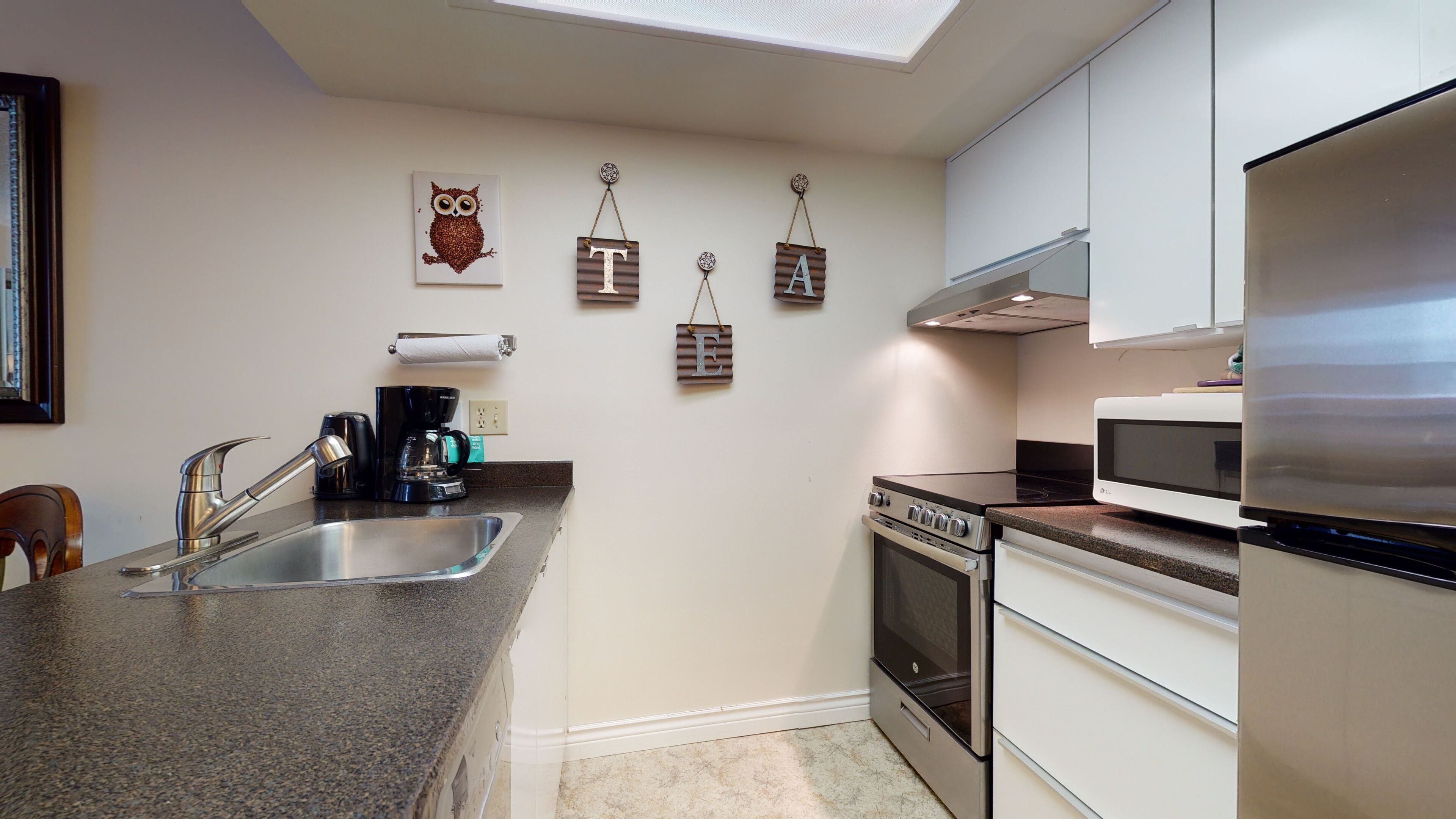 A modern kitchen with a stainless steel refrigerator, a built-in oven, and a microwave. There is a coffee maker on the counter, and the wall features decorative wooden letters and an owl painting.
