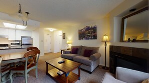 A living room with a sofa, a coffee table, a dining table with chairs, a kitchen area, and a fireplace.